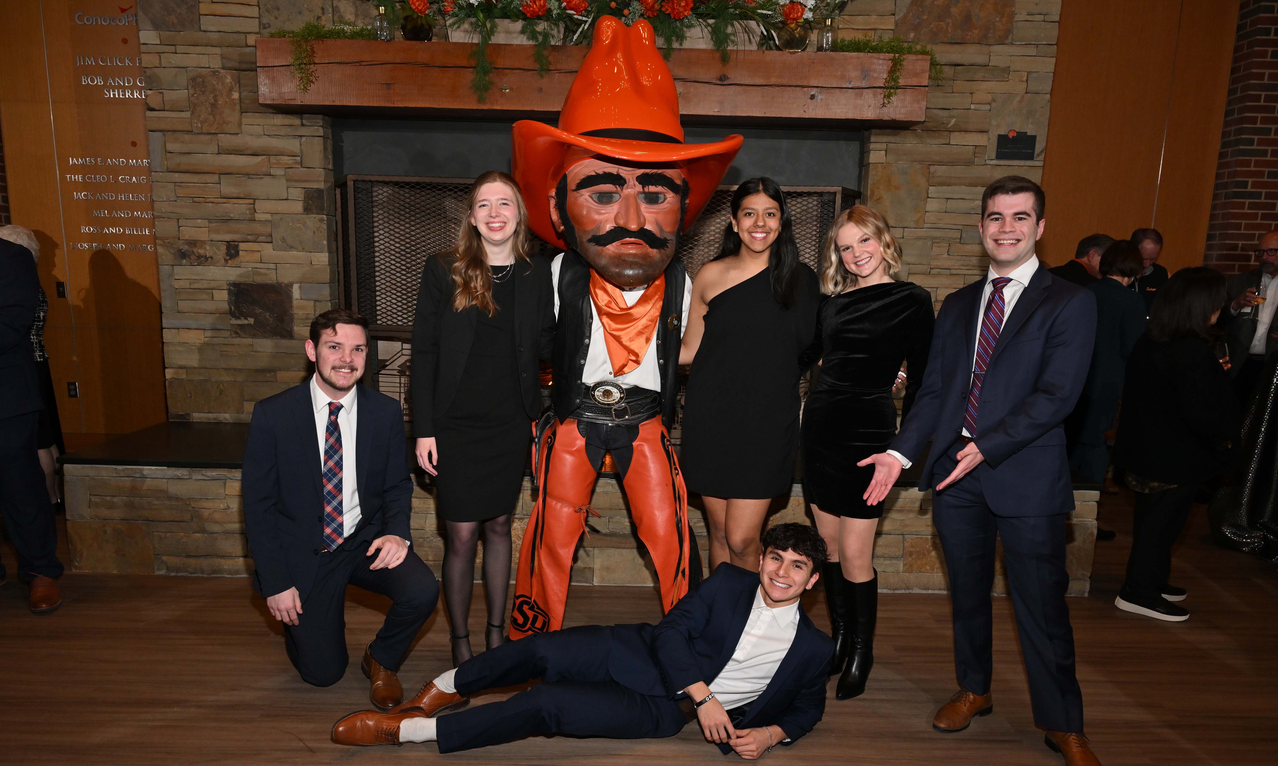 cloudofhonor-25-gallery-4.jpg Group of students in formal attire posing for a funny photo with the Pistol Pete mascot. One student laying on the groud propped up by his arm and smiling.