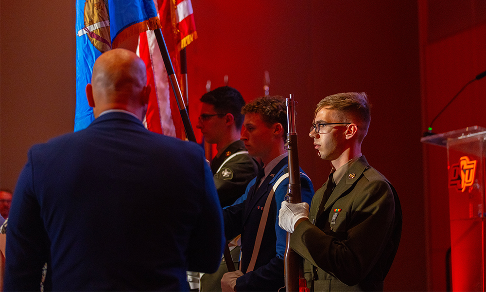 The honor guard presents the U.S. and Oklahoma state flags while a third guard holds a rifle.